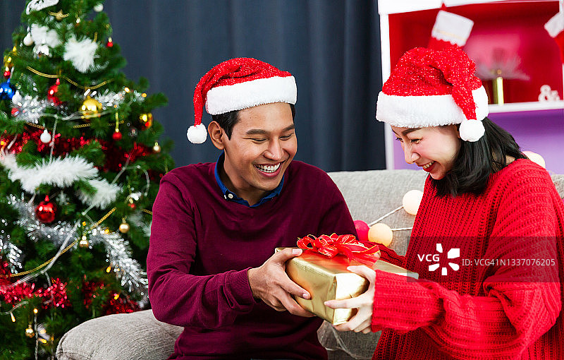 圣诞节快乐，年轻情侣互赠圣诞礼物，浪漫情侣交换礼品盒，情侣戴红色圣诞帽，圣诞假日庆祝图片素材