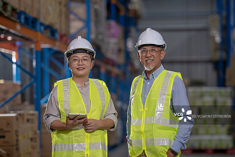 在仓库里微笑看着镜头的亚洲经理，戴着白色安全帽并拿着平板电脑图片素材