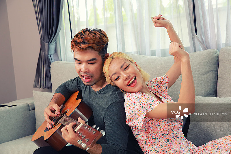 快乐的情侣在客厅的沙发上用原声吉他唱歌跳舞，人们在新冠疫情封锁期间的生活方式图片素材