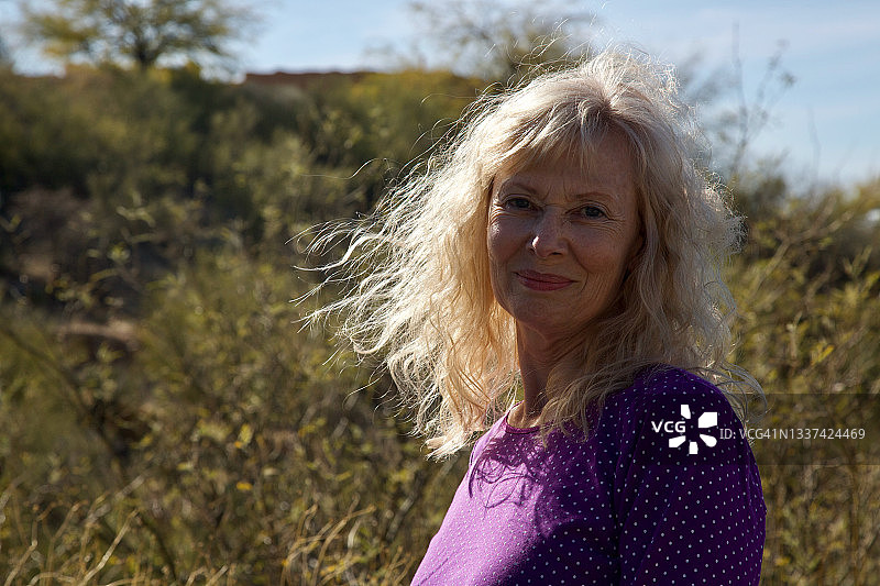 在沙漠中，金发被风吹拂，穿着紫色波点衬衫微笑的女人图片素材