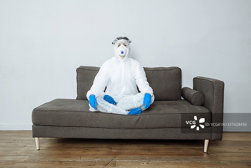 穿着防护 PPE 服、医用口罩、眼镜和手套的医生以莲花姿势坐在沙发上看着镜头图片素材