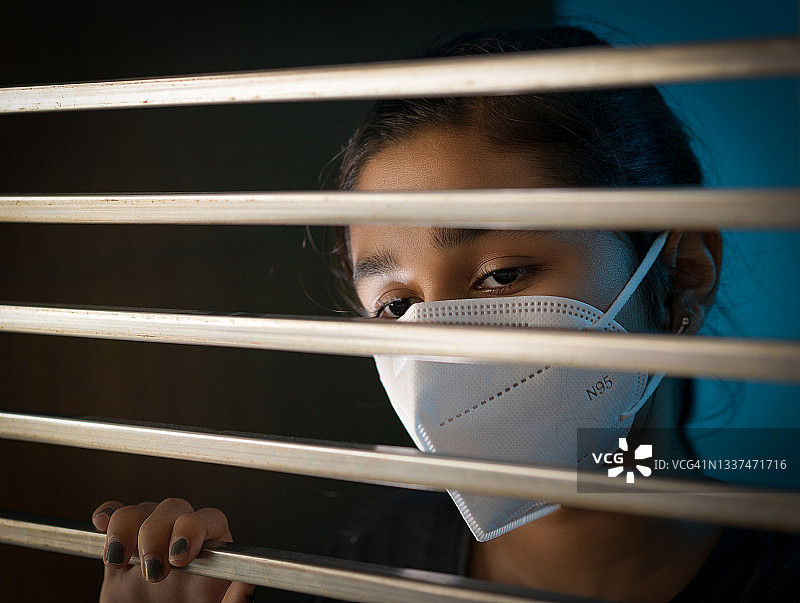 悲伤的女孩戴着防护口罩站在窗边，在隔离中沉思图片素材