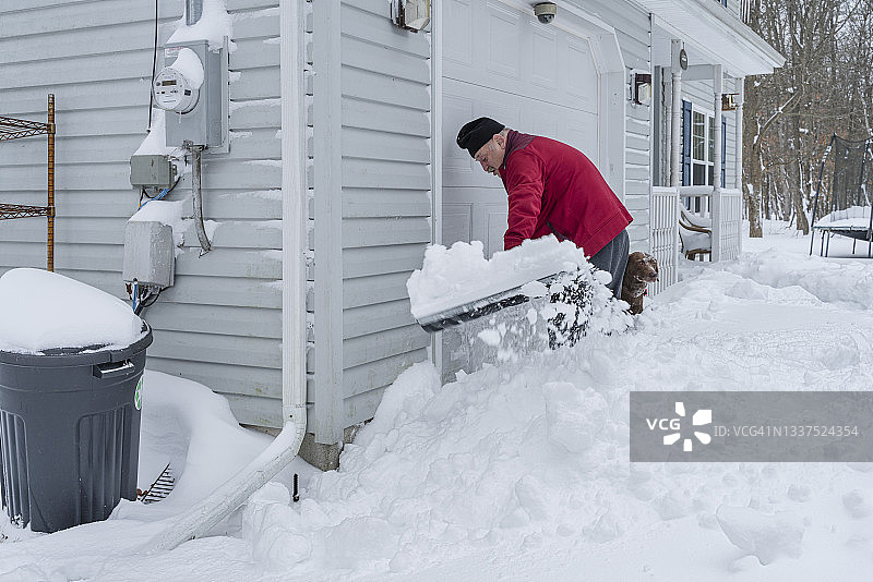年长男人在他的车库前铲雪，他的狗陪伴着图片素材
