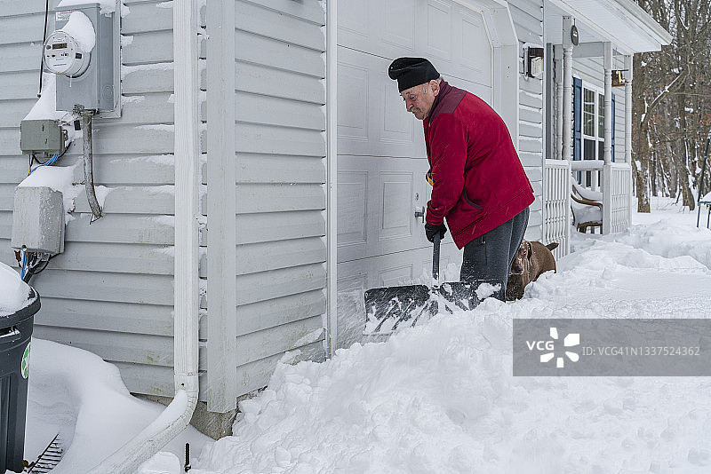 年长男人在他的车库前铲雪，他的狗陪伴着图片素材