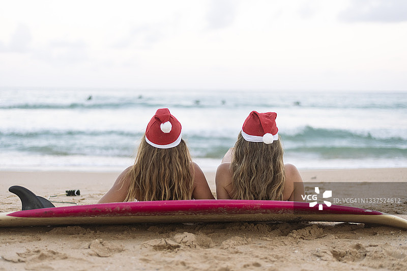 两个戴着圣诞帽的年轻女子在圣诞节期间在海滩上晒日光浴图片素材