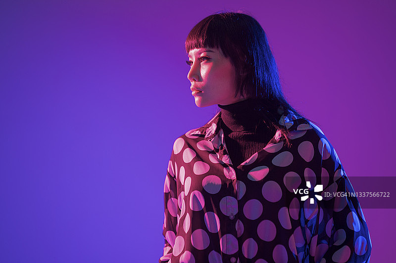 演播室中身穿圆点衬衫摆姿势的亚裔华裔女性时尚模特，霓虹蓝紫色灯光半身像图片素材