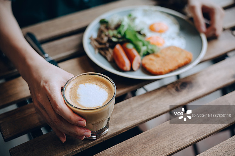 年轻女子坐在木桌旁，喝着咖啡，吃着煎蛋、土豆饼、香肠和新鲜沙拉早餐的特写图片素材