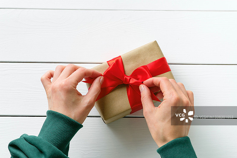 穿着绿色毛衣的女孩在木制背景或桌子上拿着一个用明亮丝带系着的精美惊喜盒子。一位女人在她的节日里包装礼物或打开礼物，系上蝴蝶结。圣诞快乐的概念。图片素材