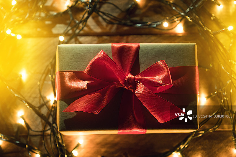 牛皮纸包装的惊喜礼物，用鲜红的丝带系着，在木桌上，背景是金色闪烁的童话灯串。在节日的前夜给予和赠送礼物。快乐圣诞节的概念图片素材