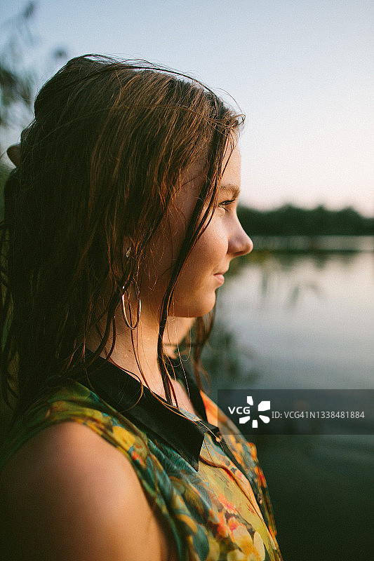湖岸边，夕阳下站在高高的草丛和水面背景前的湿发彩色连衣裙少女图片素材