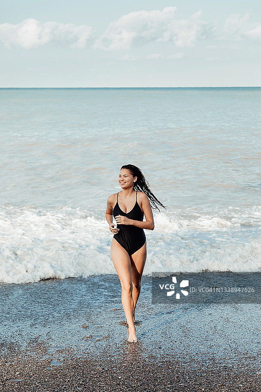 黑发年轻亚洲女孩穿着黑色泳衣从海里跑出来，侧身微笑图片素材