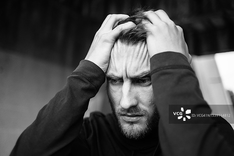 年轻的络腮胡男子黑白肖像特写，他用手抱着头，看向一边。他的面部表情充满了绝望、痛苦、后悔和愤怒。图片素材
