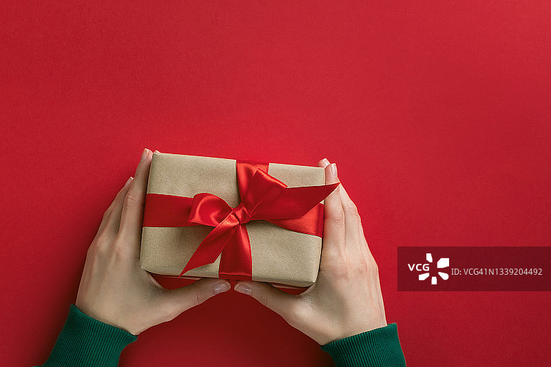 身穿绿色毛衣的女孩拿着一个漂亮的盒子，盒子上系着一条鲜艳的丝带，背景是红色。女人在节日里打开礼物。快乐圣诞节的概念图片素材