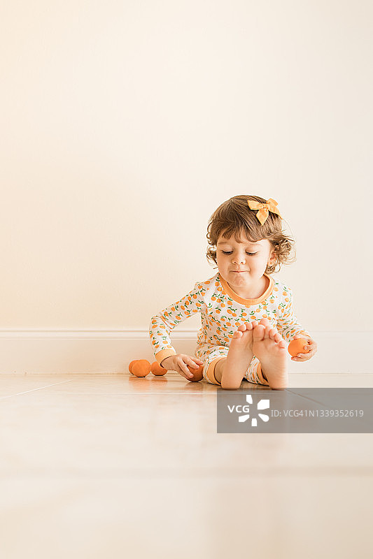两岁幼儿女孩坐在地板上玩耍和吃橘子，穿着橙色睡衣，上面有橙色橘子，并在2021年9月在家中戴着橙色发弓图片素材