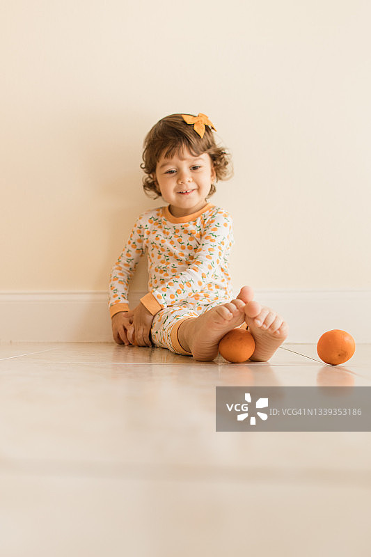 两岁幼儿女孩坐在地板上玩耍和吃橘子，穿着橙色睡衣，上面有橙色橘子，并在2021年9月在家中戴着橙色发弓图片素材