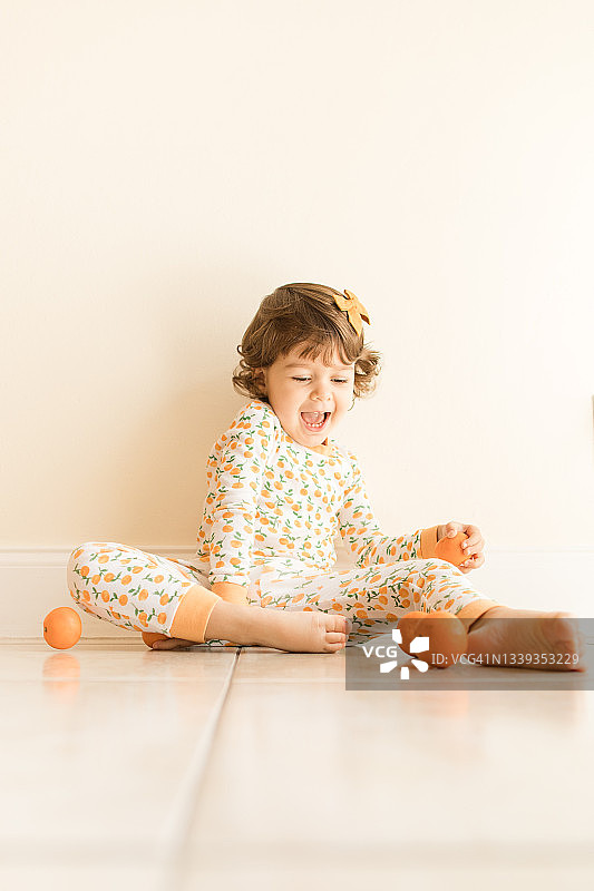 两岁幼儿女孩坐在地板上玩耍和吃橘子，穿着橙色睡衣，上面有橙色橘子，并在2021年9月在家中戴着橙色发弓图片素材
