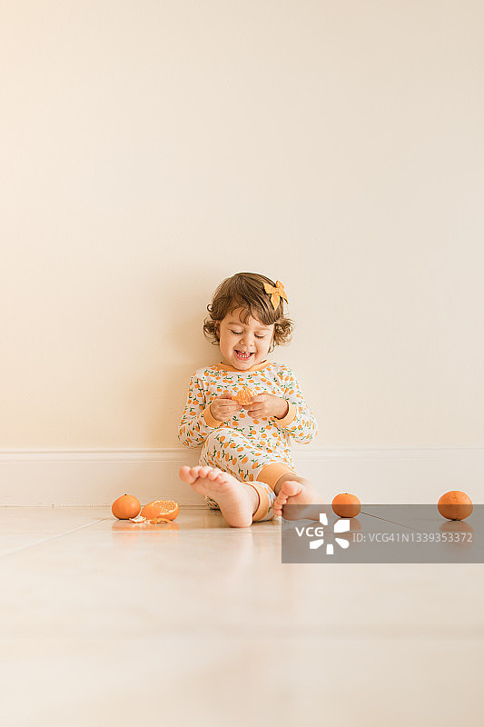 两岁幼儿女孩坐在地板上玩耍和吃橘子，穿着橙色睡衣，上面有橙色橘子，并在2021年9月在家中戴着橙色发弓图片素材