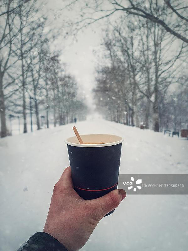 在寒冷的雪天早晨，手持一杯新鲜热咖啡在冬季公园里的人。可回收纸杯配有木制咖啡搅拌器。在摩尔多瓦基希讷乌玫瑰谷享用饮品（竖版特写生活方式照）图片素材