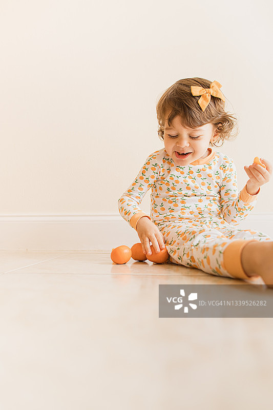 两岁幼儿女孩坐在地板上玩耍和吃橘子，穿着橙色睡衣，上面有橙色橘子，并在2021年9月在家中戴着橙色发弓图片素材