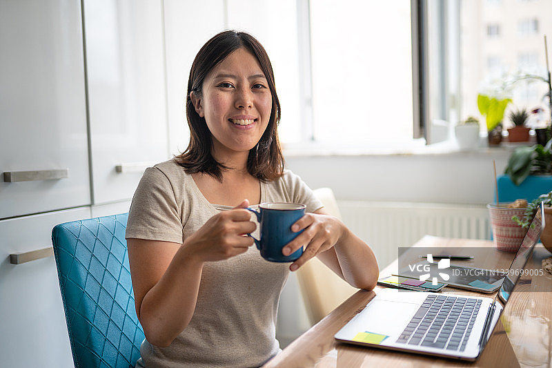 年轻女人在家一边用笔记本电脑工作一边喝咖啡对着镜头微笑图片素材