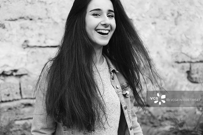 站在砖墙背景前看着镜头大笑的毛衣牛仔外套长发美女。黑白摄影，正面特写图片素材