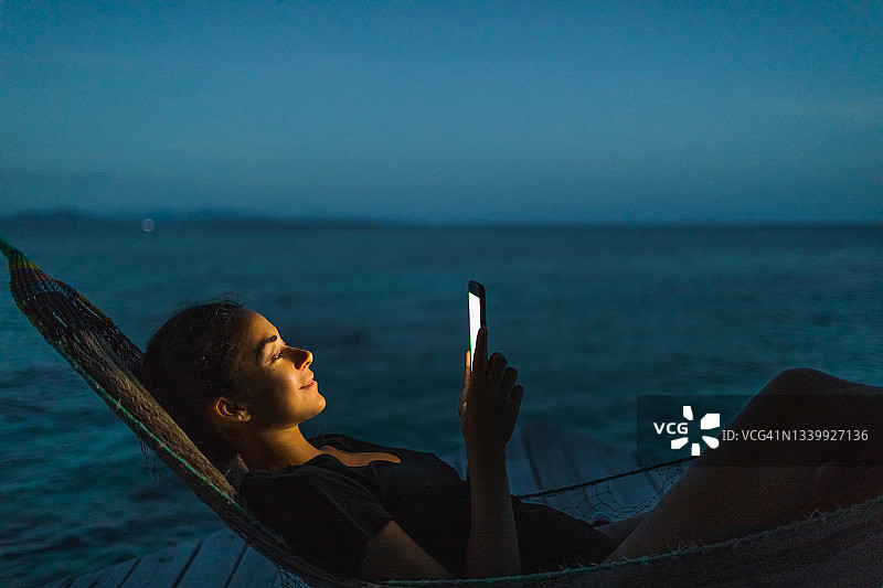 夜晚，女人在吊床上放松地发短信，与朋友在线聊天图片素材