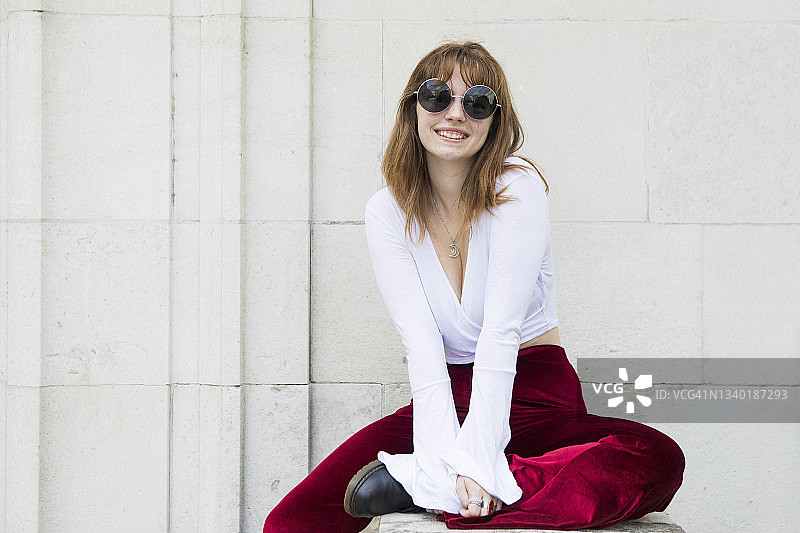 时尚的年轻女人坐在石墙边，看着镜头微笑图片素材