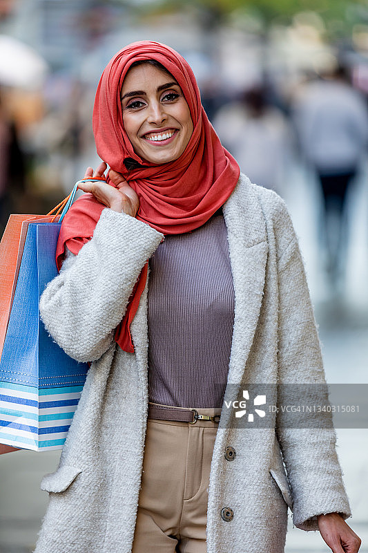 戴着红色头巾的穆斯林妇女肩上挎着彩色购物袋，走在欧洲老城的街道上。城市购物和折扣概念图片素材