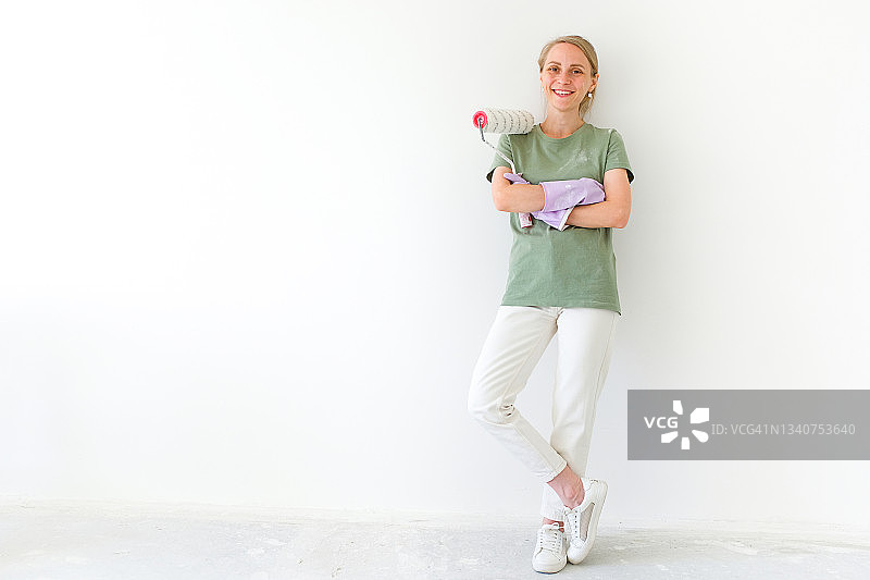 一位快乐、微笑的女建筑工人或油漆工拿着一个滚筒，滚筒上溅满了油漆，她靠在白墙上，看着镜头，用于家庭和专业建筑和维修的工具，建筑行业图片素材