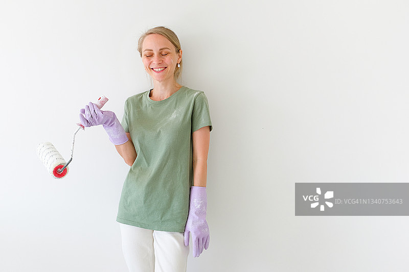 一位快乐、微笑的女建筑工人或油漆工拿着一个滚筒，滚筒上溅满了油漆，她靠在白墙上，看着镜头，用于家庭和专业建筑和维修的工具，建筑行业图片素材