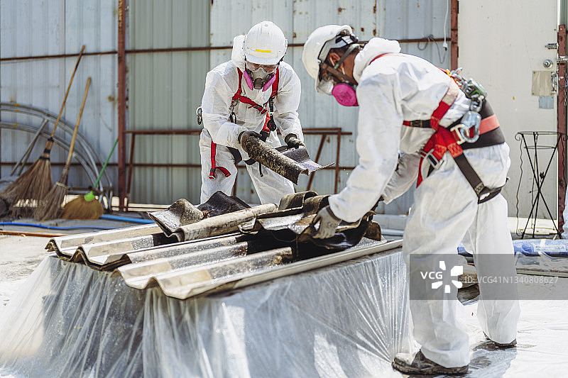 安全是我们的首要任务。工人在使用石棉屋顶瓦时穿着全身防护服。图片素材