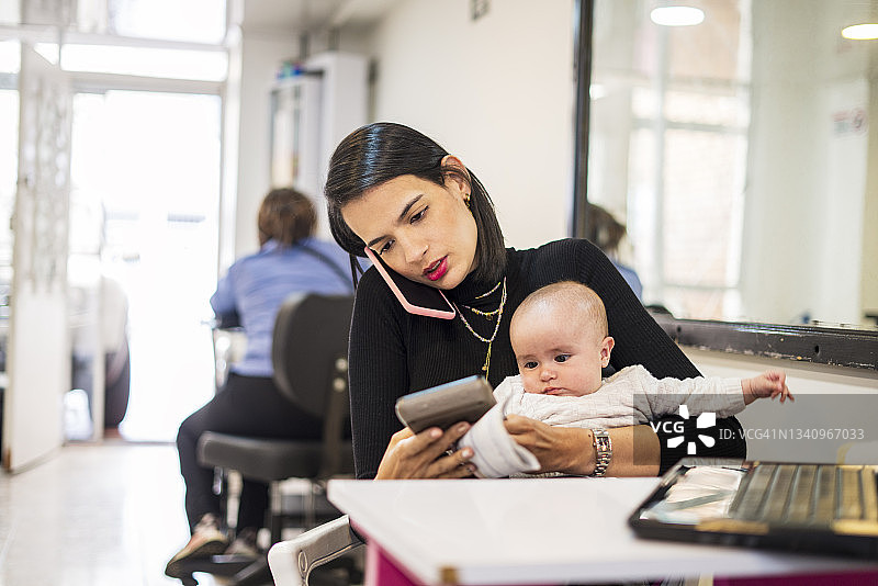 美发店经理抱着孩子工作，接电话并在笔记本电脑上打字图片素材