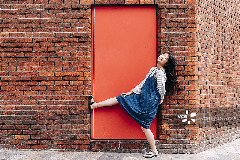 快乐的女人靠在砖墙前的红门旁图片素材