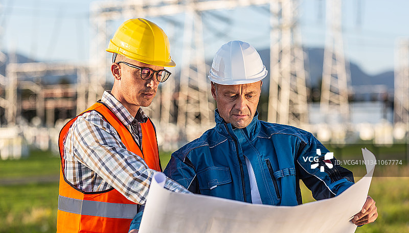 工头和戴着安全帽的工人在夏日里查看蓝图计划，背景是配电中心图片素材
