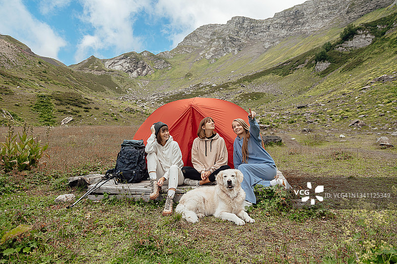 三个年轻欧洲女人带着金毛猎犬坐在山间露营帐篷旁图片素材