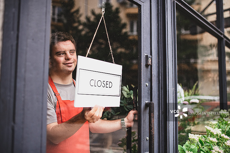 患有唐氏综合征的年轻男花店员工在花店内拿着停止营业的牌子图片素材