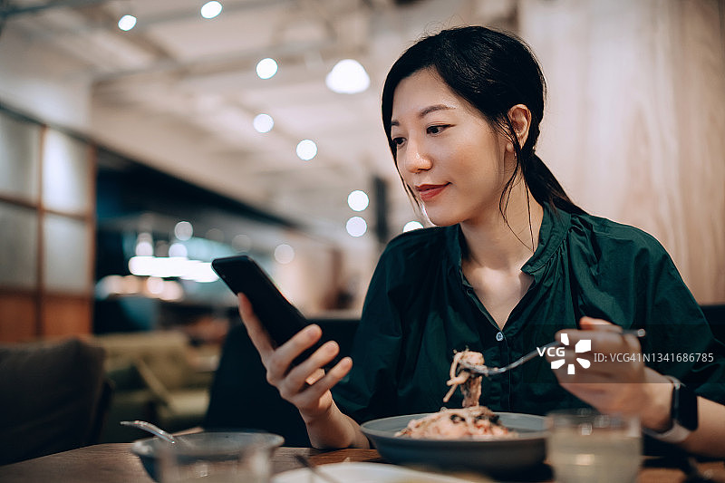年轻的亚洲女性在餐厅午餐时用智能手机发短信，享用新鲜的意大利面。独自享受轻松的午休。生活方式与科技图片素材
