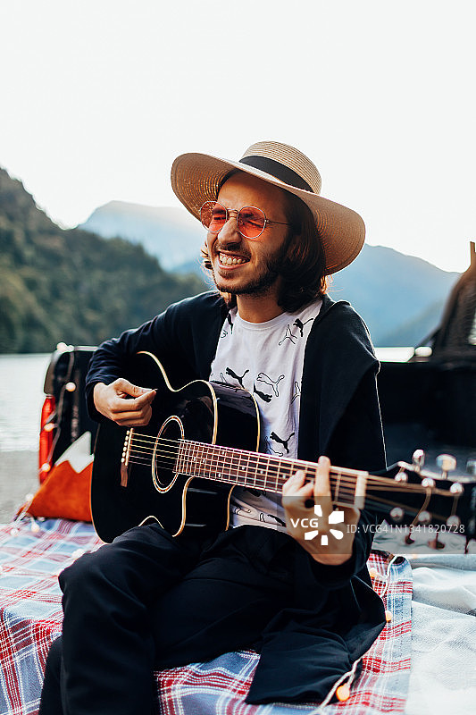 戴着帽子和粉色太阳镜的年轻欧洲男子在日落时坐在皮卡车后部弹吉他唱歌图片素材