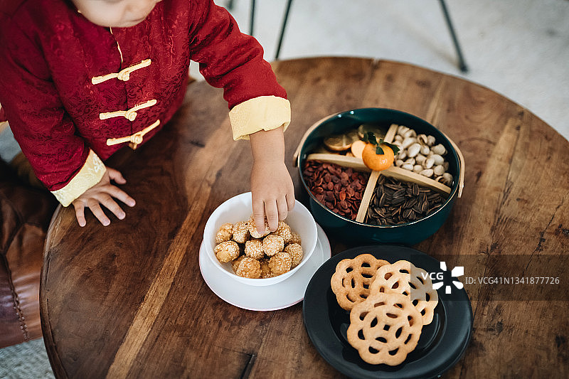 可爱的小亚裔男孩穿着中国服装，从木桌上抓取中国新年传统小吃图片素材