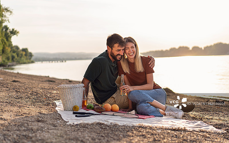 一对拥抱在一起的年轻夫妇在河边享受宁静而随意的野餐，喝了几杯啤酒后开怀大笑图片素材