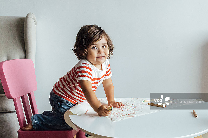 穿着红白条纹衬衫的小男孩坐在桌旁，一边看着镜头，一边用彩色铅笔在客厅里画画图片素材