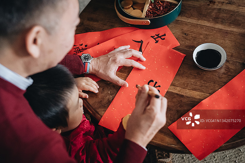 爷爷在家练习中国书法，为春节写挥春（吉祥话）并教孙子写对联图片素材