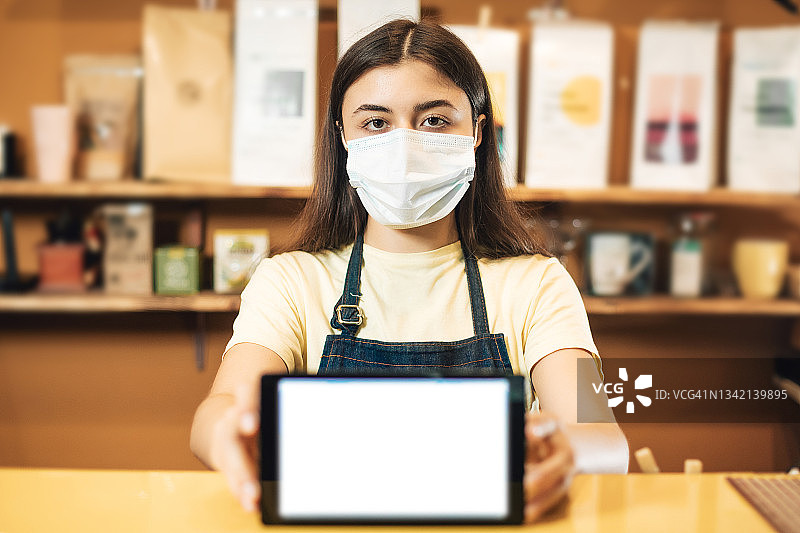戴防护口罩的年轻咖啡师在咖啡馆工作，手持平板电脑图片素材