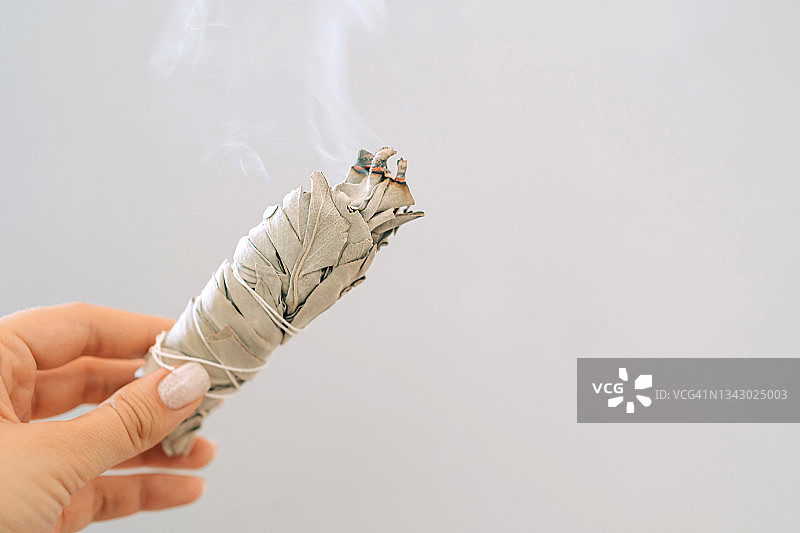 女人手持燃烧的白色鼠尾草，灰色背景，房间烟熏仪式图片素材
