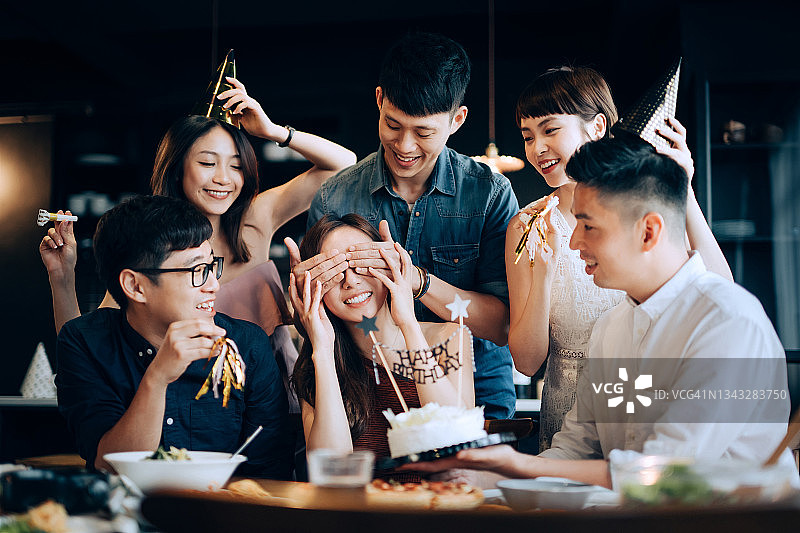 一群快乐的年轻朋友一起庆祝惊喜生日派对。朋友们在派对上欢呼雀跃，用生日蛋糕给她带来惊喜，其中一个男人捂住了生日女孩的眼睛图片素材
