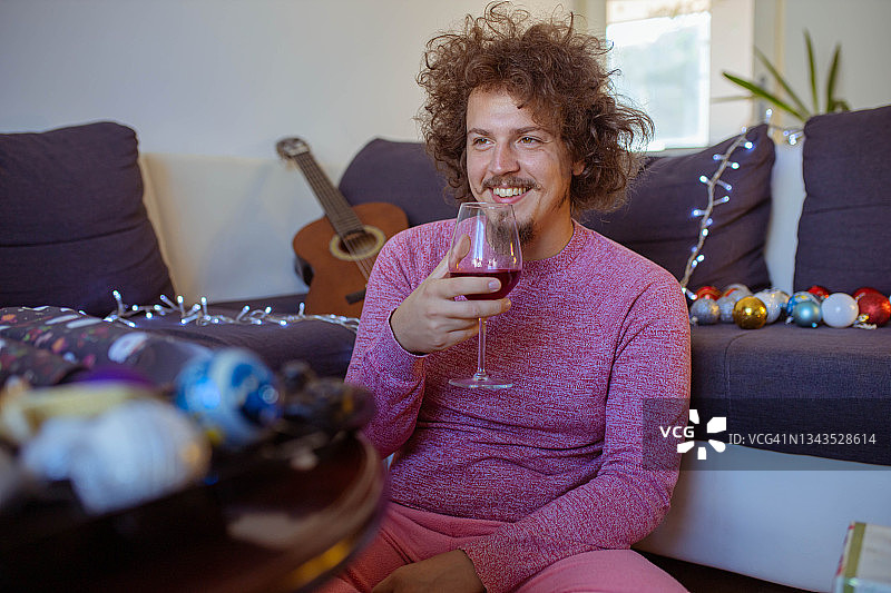 快乐的年轻男子穿着睡衣，独自一人在假期里看电影，喝红酒图片素材