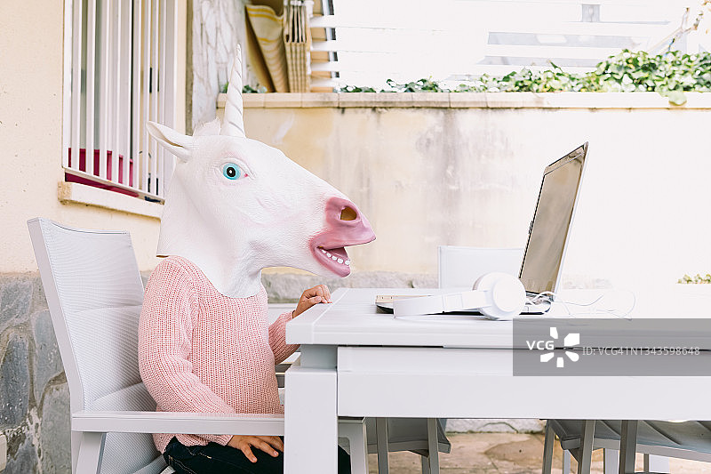 孩子戴着独角兽乳胶面具，穿着粉色毛衣，坐在露台的笔记本电脑前。在线远程教育、童年、科技与超现实主义概念。图片素材