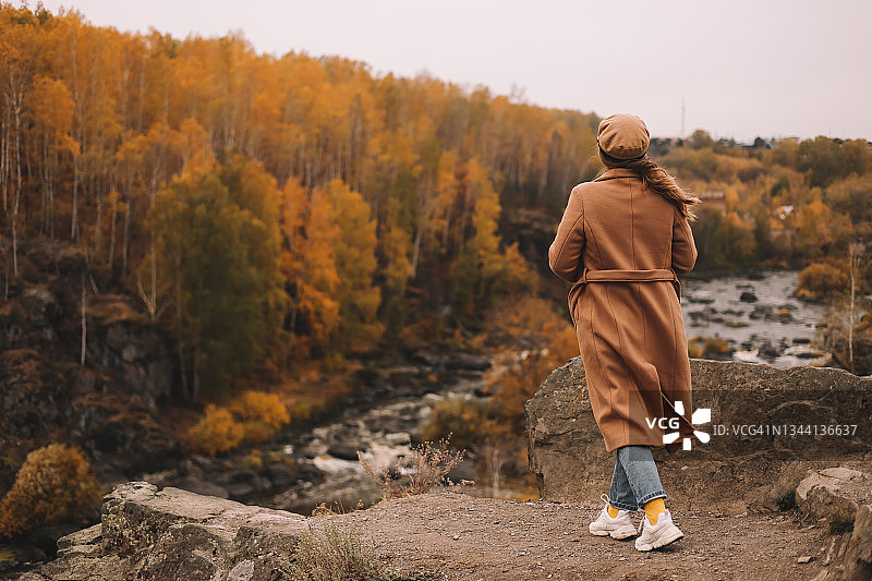 身穿休闲暖外套和帽子的快乐年轻女子在秋季户外旅行，享受在秋季森林中沿着河流和山脉漫步的孤独图片素材