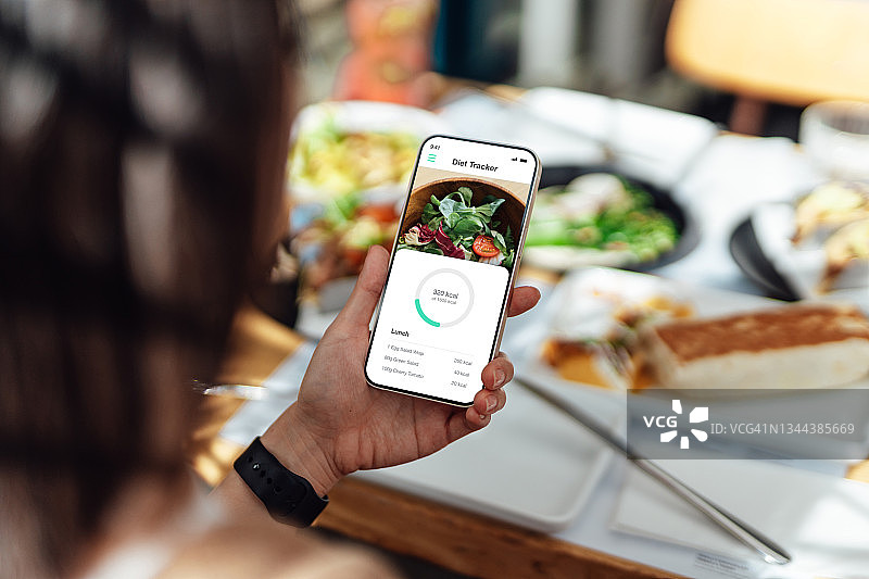 年轻女子在户外用餐时用手机应用程序查看卡路里摄入量（肩上视角）图片素材