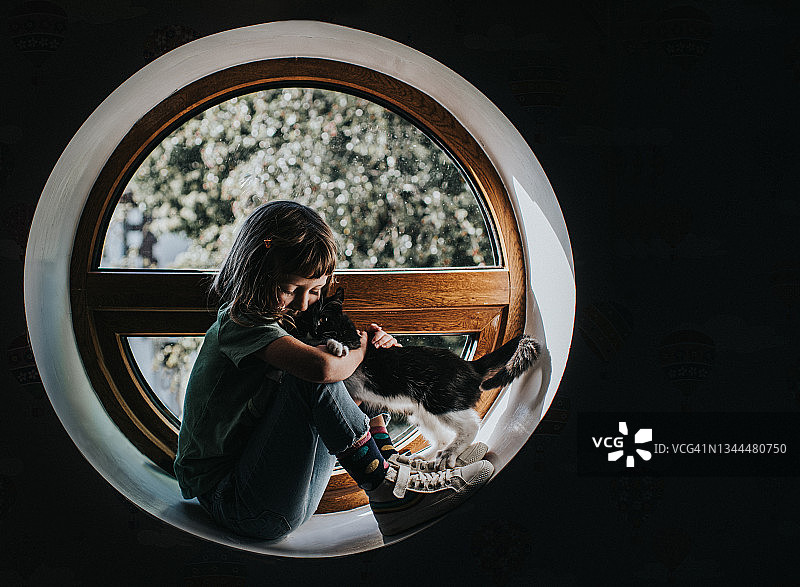 一个年轻女孩紧紧地抱着一只小猫，他们坐在圆形窗户旁的窗台上图片素材